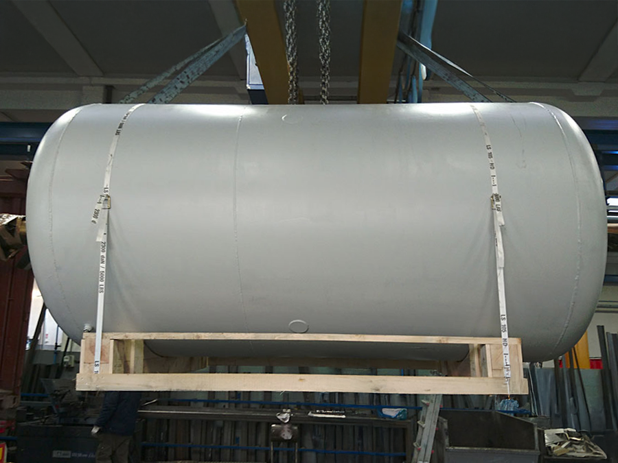 Un grande serbatoio metallico cilindrico viene sollevato all'interno di un impianto industriale utilizzando cinghie e una gru. Il serbatoio è sostenuto nella parte inferiore da un telaio di legno.