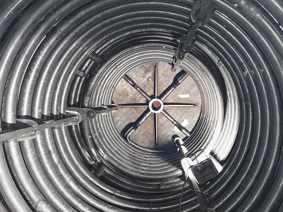 Vista dall'alto di una grande struttura metallica cilindrica con tubi o cavi arrotolati intorno alla parete interna e un asse centrale con supporti metallici incrociati. Sul lato interno sono fissati attrezzi o macchinari.