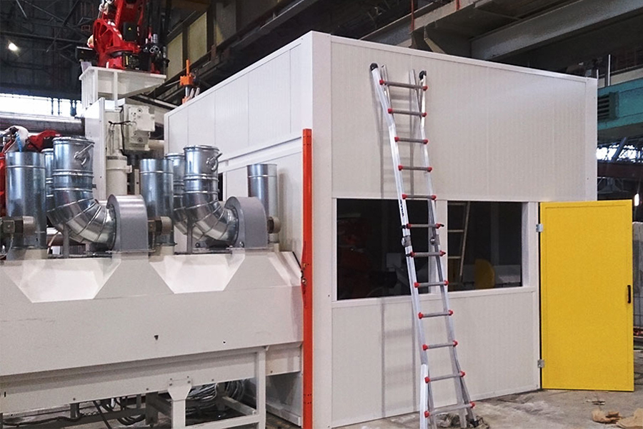 Un macchinario industriale bianco con grandi tubi metallici è posizionato accanto a una stanza modulare bianca, con una porta gialla e una finestra. Una scala alta è appoggiata all'edificio. La scena si svolge all'interno di una fabbrica.