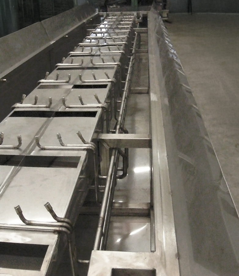 Grande sistema di trasporto industriale in metallo con ganci verticali uniformemente distanziati e vassoi rettangolari, situato all'interno su un pavimento in cemento, probabilmente utilizzato per la lavorazione o la produzione di alimenti.