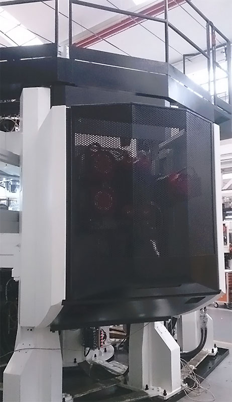 Un grande macchinario industriale con una copertura di sicurezza nera perforata e pannelli esterni bianchi si trova in un ambiente industriale, con ringhiere metalliche e tubi visibili sullo sfondo.