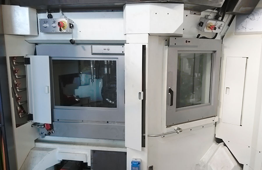 Macchina CNC industriale con ampie finestre e pannelli di controllo, con pulsanti di arresto di emergenza e vari interruttori, all'interno di uno spazio di lavoro pulito e chiuso.