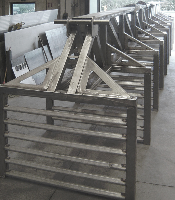 Diverse grandi strutture metalliche con supporti inclinati e barre orizzontali sono allineate all'interno, probabilmente in un'officina o in un ambiente industriale, con lamiere e attrezzature visibili sullo sfondo.