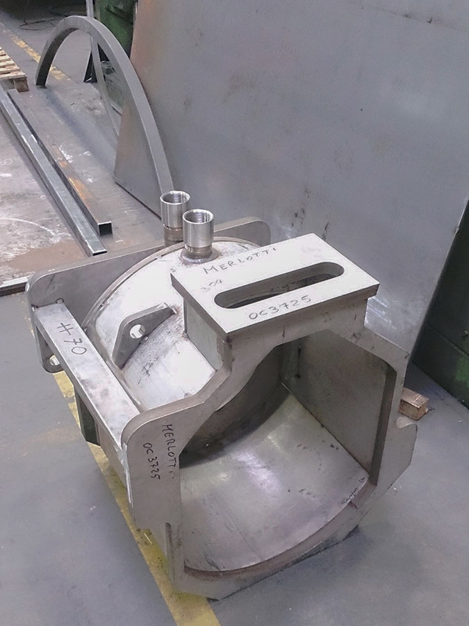 Un grande componente industriale metallico con caratteristiche cilindriche e rettangolari è collocato sul pavimento di una fabbrica. Il pezzo presenta etichette e due tubi metallici sulla parte superiore e sembra far parte di un macchinario o di un'attrezzatura pesante.