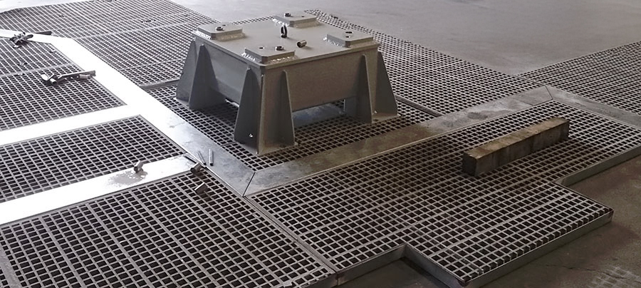 Un pavimento metallico con grate a griglia, una struttura metallica rettangolare rialzata al centro e un blocco di cemento rettangolare adagiato sulla grata vicina.