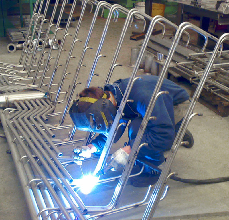 Una persona che indossa un equipaggiamento protettivo è accovacciata a saldare tubi metallici in un'officina. Sul punto di saldatura sono visibili scintille luminose e davanti a loro sono disposti diversi tubi metallici curvi.