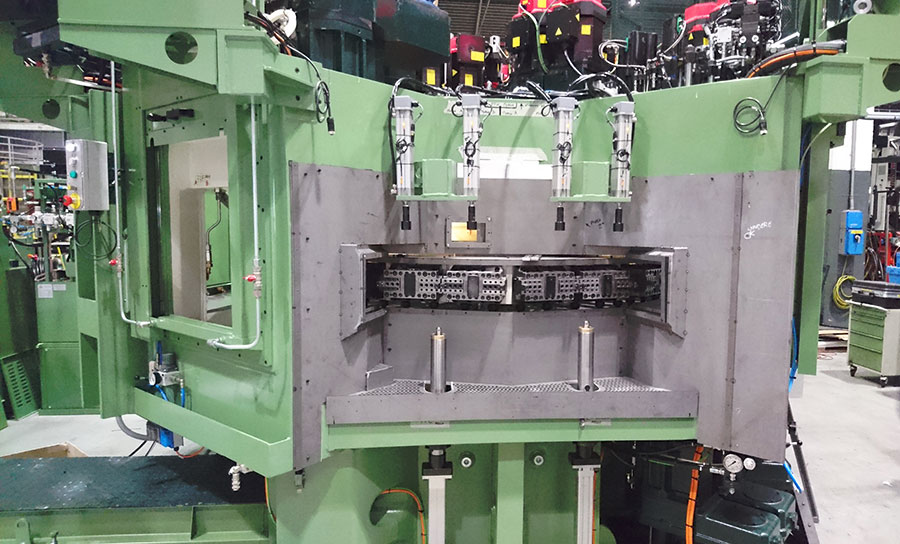 Una grande macchina industriale con un telaio verde, componenti metallici e diverse parti cilindriche, utilizzata per processi di produzione o lavorazione, è allestita all'interno di una fabbrica. In alto sono visibili i cavi e le unità di controllo.