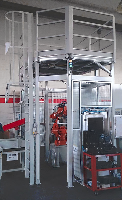 Una postazione industriale robotizzata con un braccio robotico rosso all'interno di un'area recintata, una piattaforma metallica sovrastante con ringhiera e scala, varie attrezzature e un carrello porta attrezzi rosso nelle vicinanze.