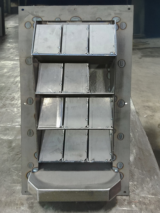 Vista ravvicinata di una presa d'aria industriale metallica con lame angolate, progettata per il controllo del flusso d'aria, montata su un telaio metallico rettangolare in un'officina.