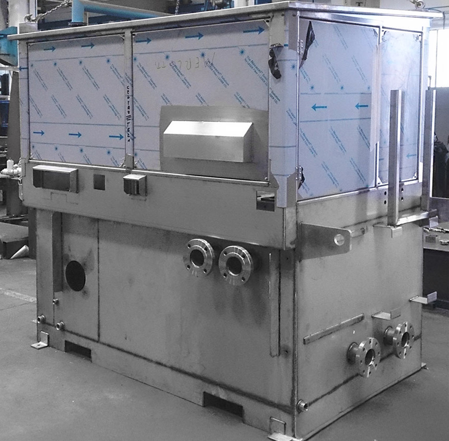 Grande macchina industriale in acciaio inox con tubi multipli, valvole e una sezione superiore rettangolare, posizionata all'interno di un'officina o di una fabbrica. I pannelli superiori sono contrassegnati da frecce blu.