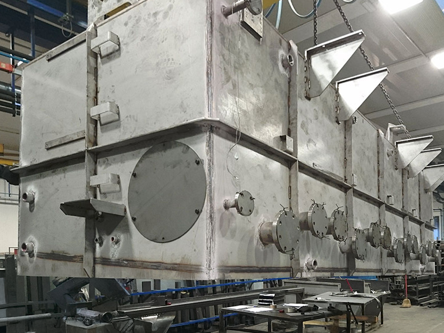 Grandi attrezzature industriali in acciaio inox con pannelli di accesso multipli, flange e raccordi sono sospese da catene in un ambiente industriale, con strutture metalliche e tavoli visibili sullo sfondo.