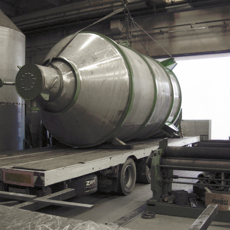 Un grande serbatoio industriale cilindrico in metallo viene caricato su un camion a pianale all'interno di una fabbrica con una gru. Il serbatoio è lucido e metallico con accenti verdi e sembra essere di recente produzione.