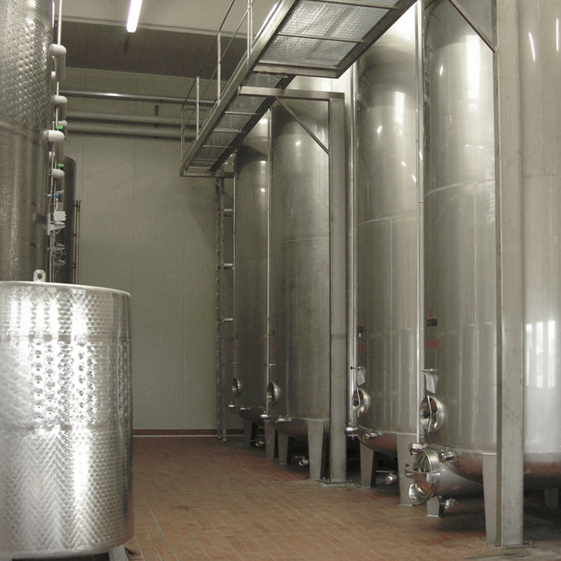 Grandi serbatoi di fermentazione in acciaio inox si trovano in una stanza industriale pulita con pavimento in mattoni e passerella sopraelevata, probabilmente utilizzata per i processi di produzione della birra o del vino.