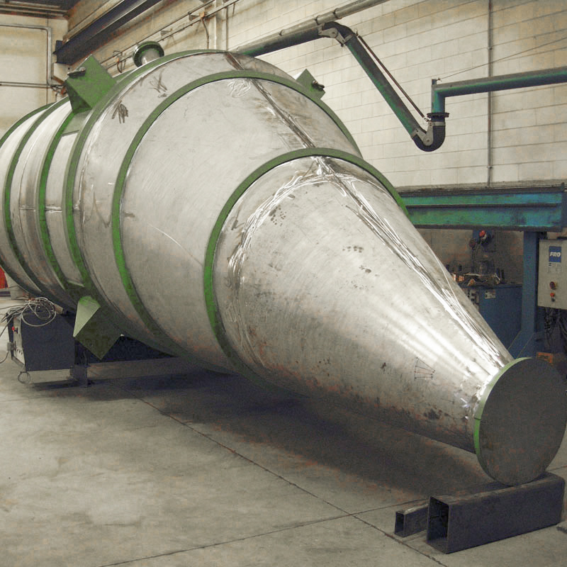 Un grande separatore industriale metallico a ciclone di forma conica si trova sul pavimento di una fabbrica con pareti piastrellate e macchinari visibili sullo sfondo.