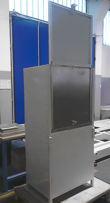 Un grande armadio industriale rettangolare in metallo con un'anta superiore aperta verticalmente, situato in un interno vicino a divisori blu e grandi finestre, con varie attrezzature e materiali sullo sfondo.