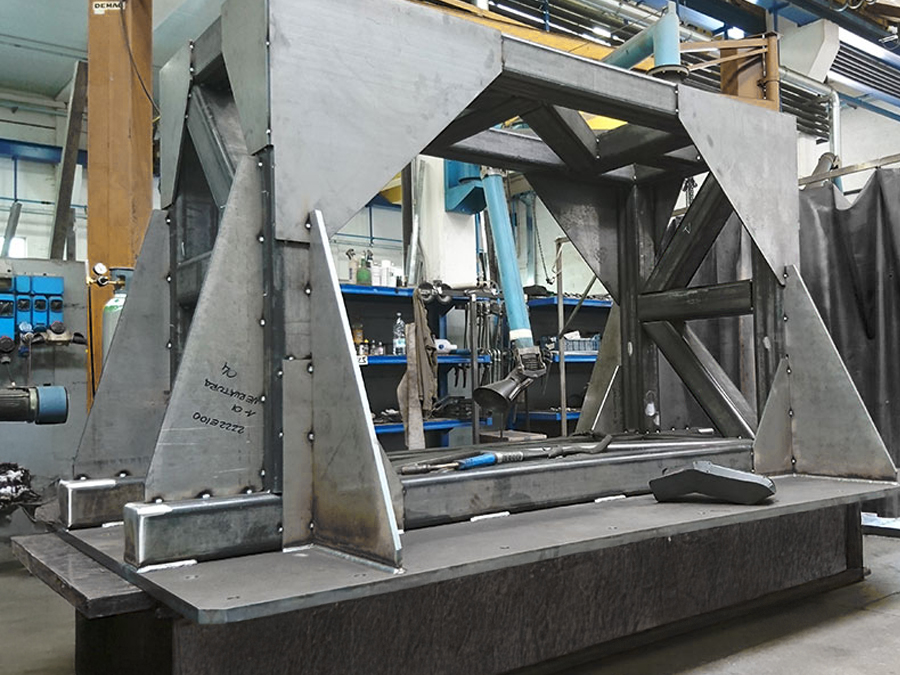 Una grande struttura rettangolare metallica si trova in un'officina industriale. Sullo sfondo sono visibili attrezzi e apparecchiature, oltre a scaffali e tubi che rivestono le pareti bianche e blu.