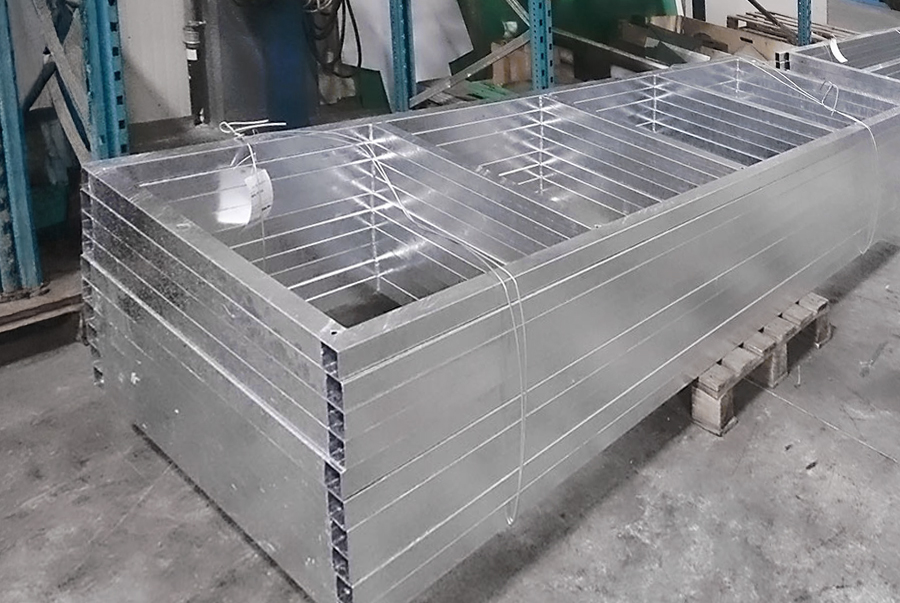 Una pila di pezzi di condotti metallici rettangolari, legati con cinghie, si trova su un pallet di legno in un magazzino industriale. Sullo sfondo sono visibili scaffali e macchinari in metallo blu.
