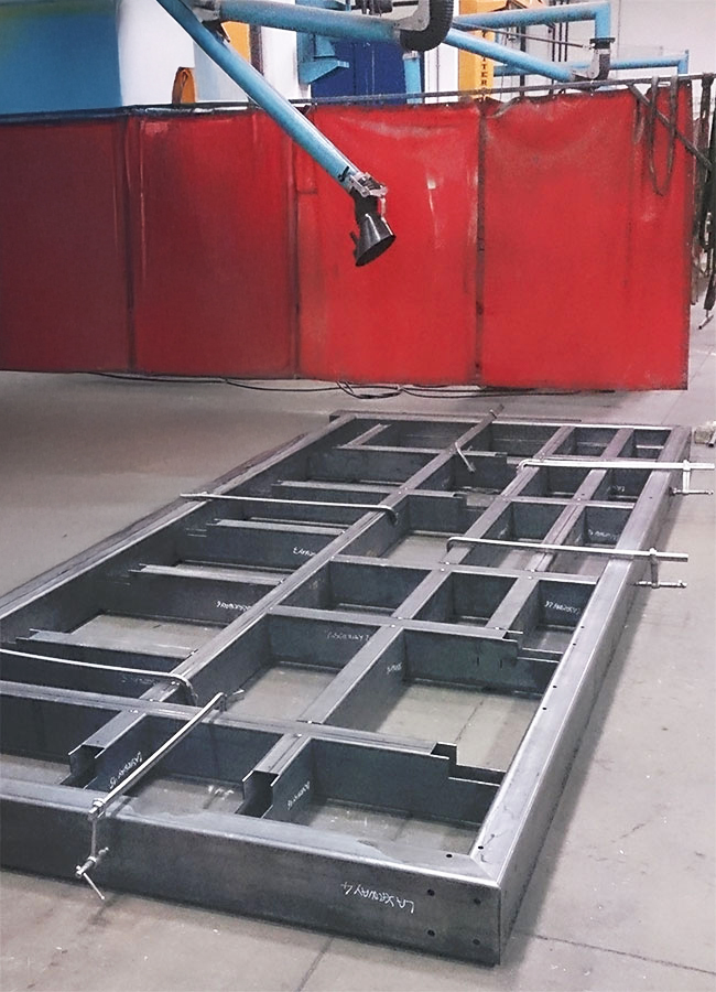 Una grande struttura metallica rettangolare con travi d'acciaio intersecate giace sul pavimento di un'officina industriale; sullo sfondo si intravedono pareti divisorie rosse e macchinari blu.