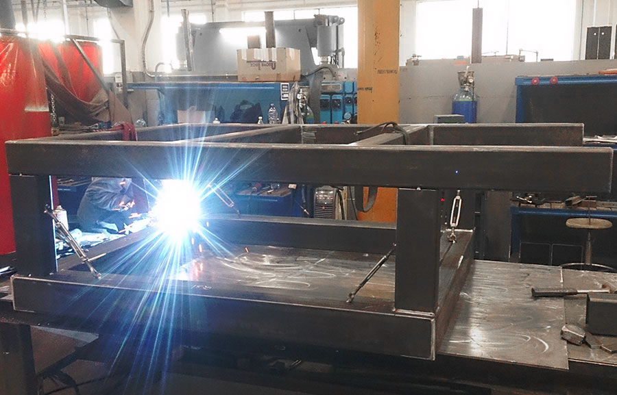 Una persona sta saldando una grande struttura metallica su un banco di lavoro in un'officina industriale, con un arco di saldatura luminoso che illumina la scena. Sullo sfondo sono visibili vari strumenti e attrezzature.