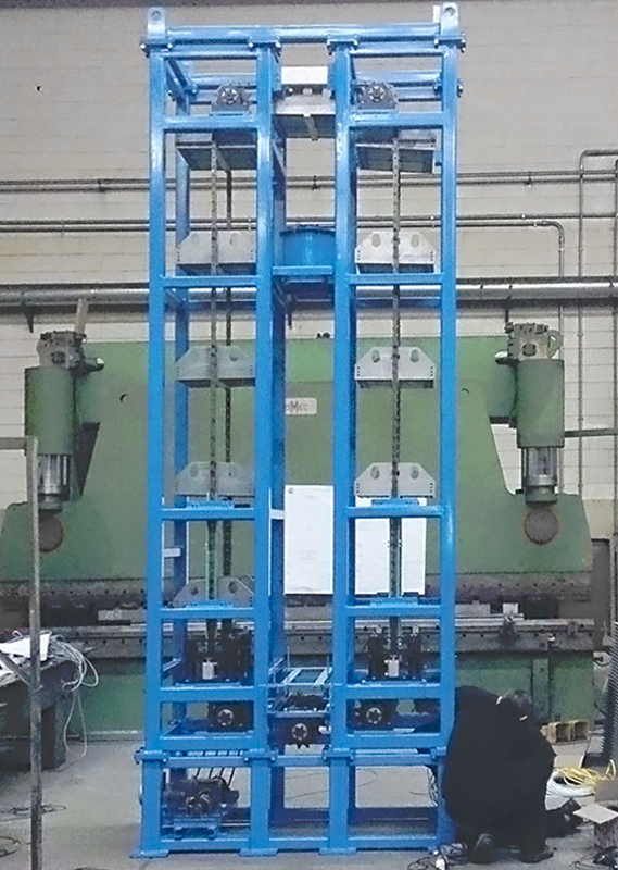 Un'alta struttura metallica industriale blu si trova in un'officina, con vari componenti meccanici e catene. Sullo sfondo sono visibili grandi macchine e utensili verdi, oltre a cavi e attrezzature sul pavimento.